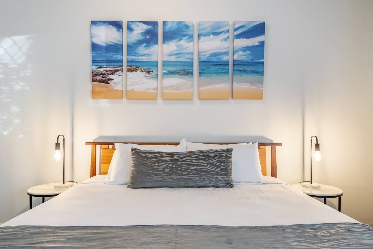 A hotel room with a bed, two bedside lamps, a nightstand, and a wall-mounted artwork of a beach scene.