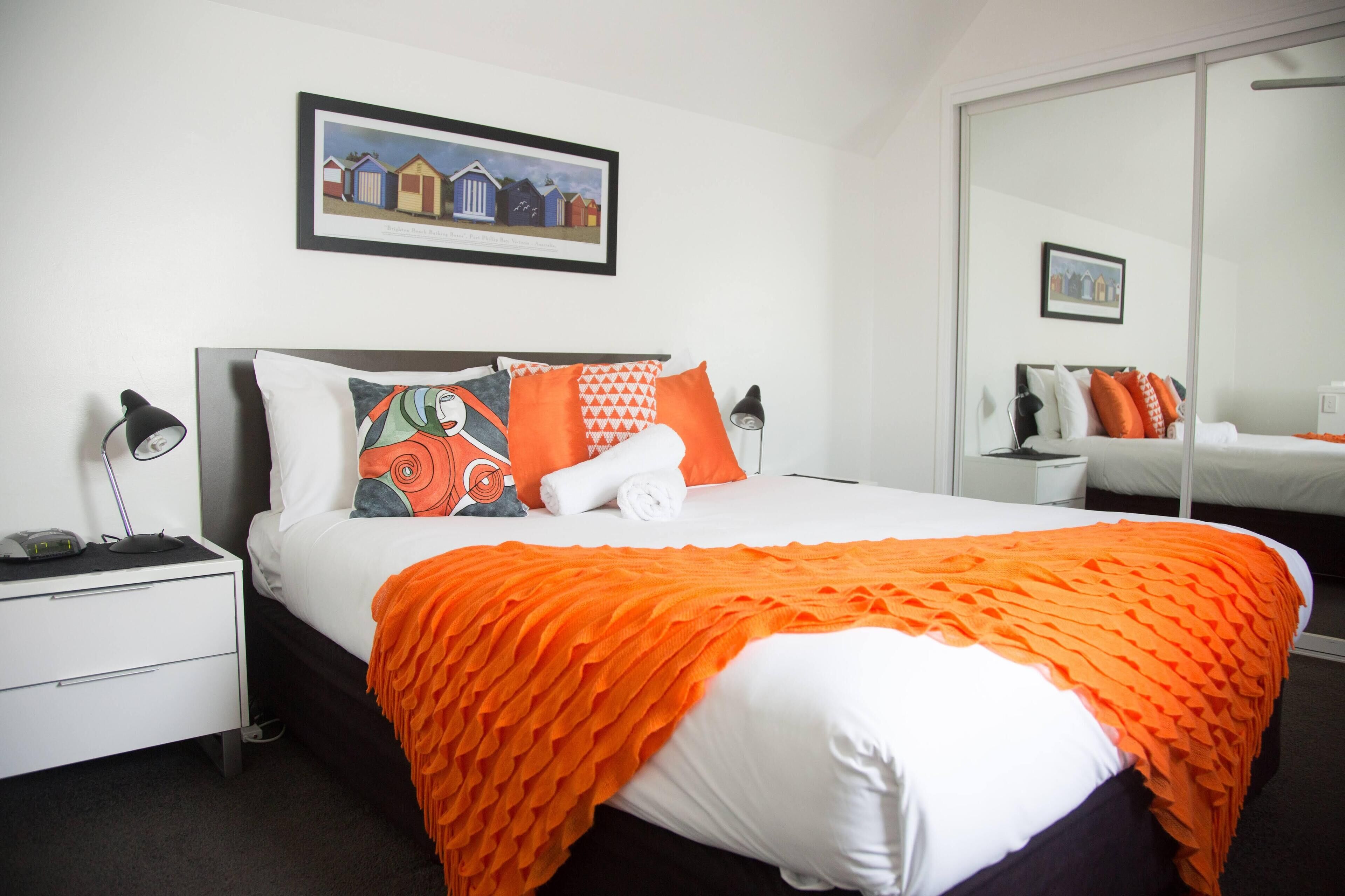 Una cama bien hecha con un edredón naranja, un almohadón blanco y un cojín colorido. Encima de la cama, en la pared, hay un cuadro enmarcado, y una mesita de noche con una lámpara junto a ella.