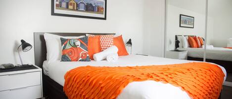 Una cama bien hecha con un edredón naranja, un almohadón blanco y un cojín colorido. Encima de la cama, en la pared, hay un cuadro enmarcado, y una mesita de noche con una lámpara junto a ella.