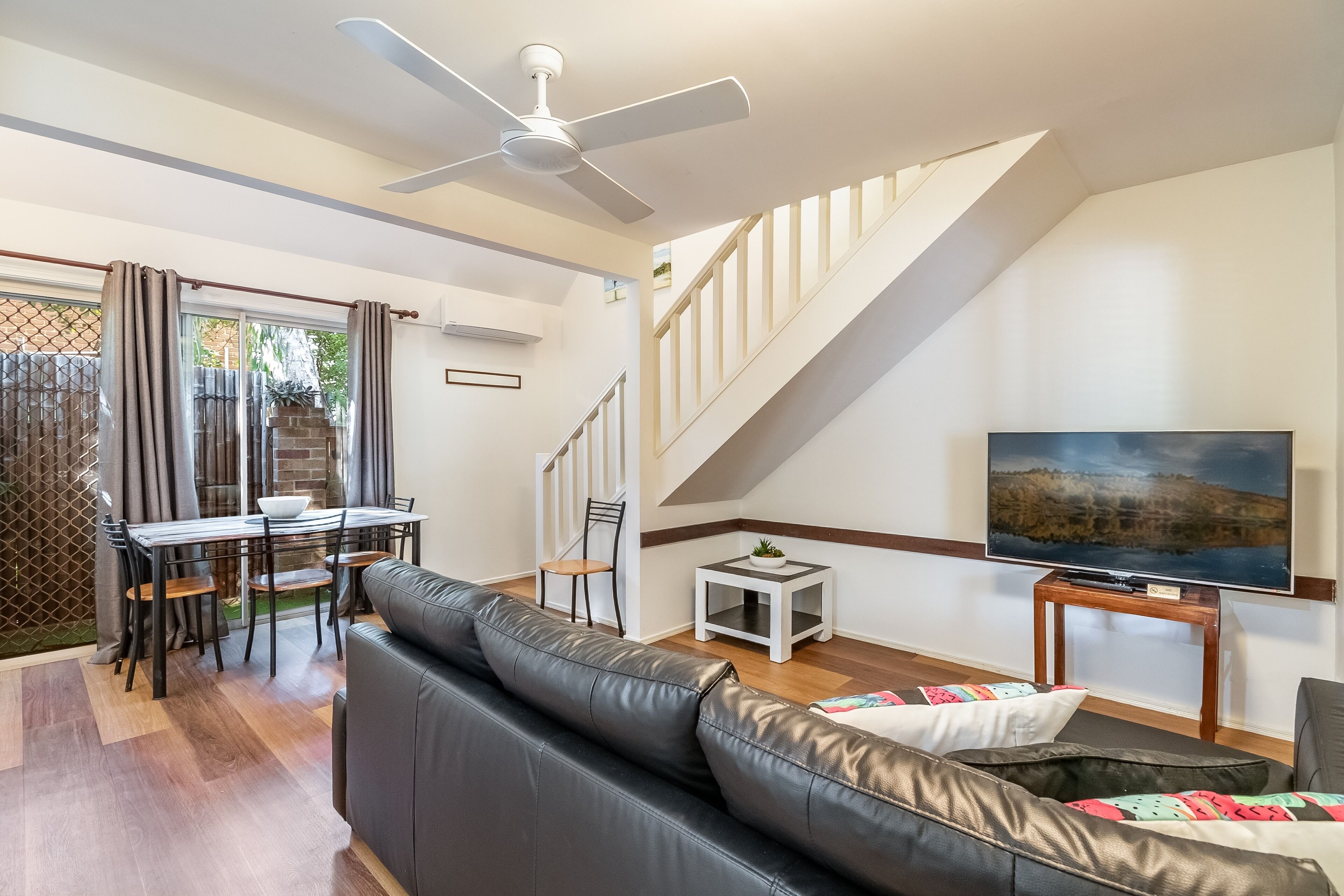 A modern living room with a black leather sofa, a dining table with chairs, a TV on a wooden stand, and a staircase with a white railing.