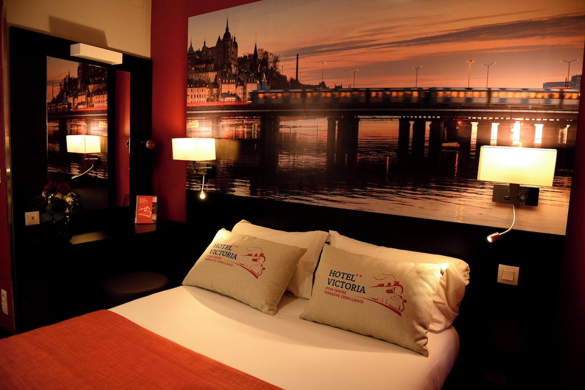A hotel room with a bed, two pillows, a nightstand with a lamp, a mirror, and a large window with a cityscape view.