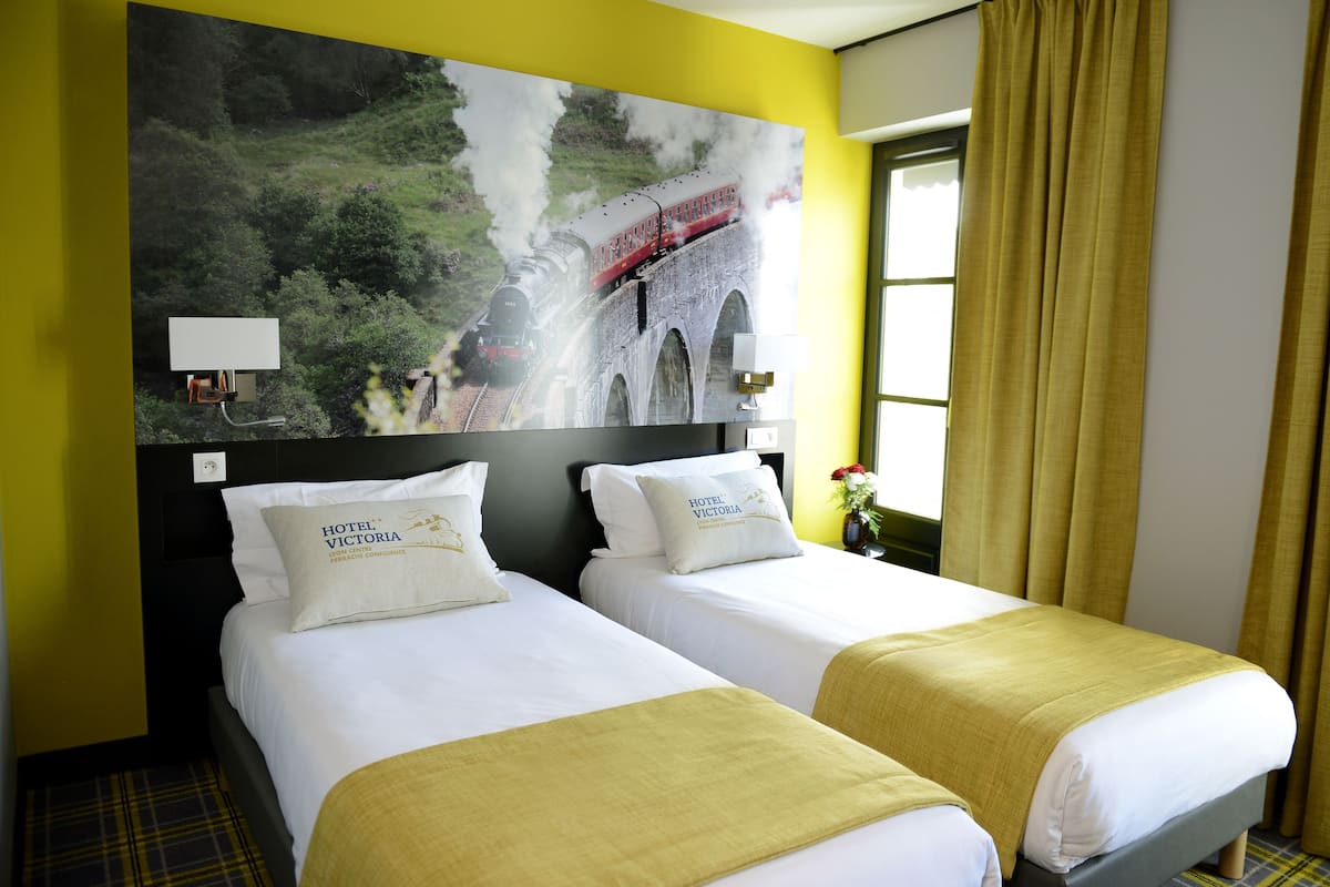 A hotel room with two beds, a large painting of a train on a bridge, and yellow curtains.