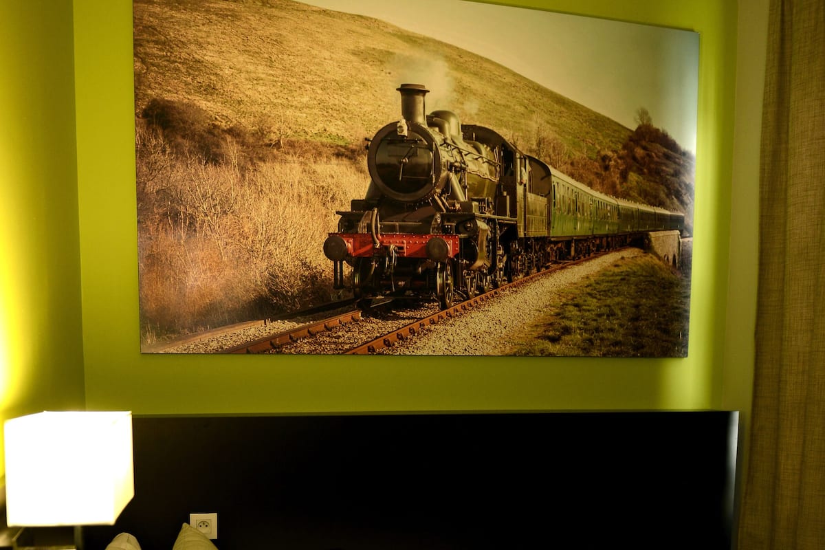 A hotel room with a bed, pillows, a bedside lamp, a potted plant, and a large painting of a train on the wall.