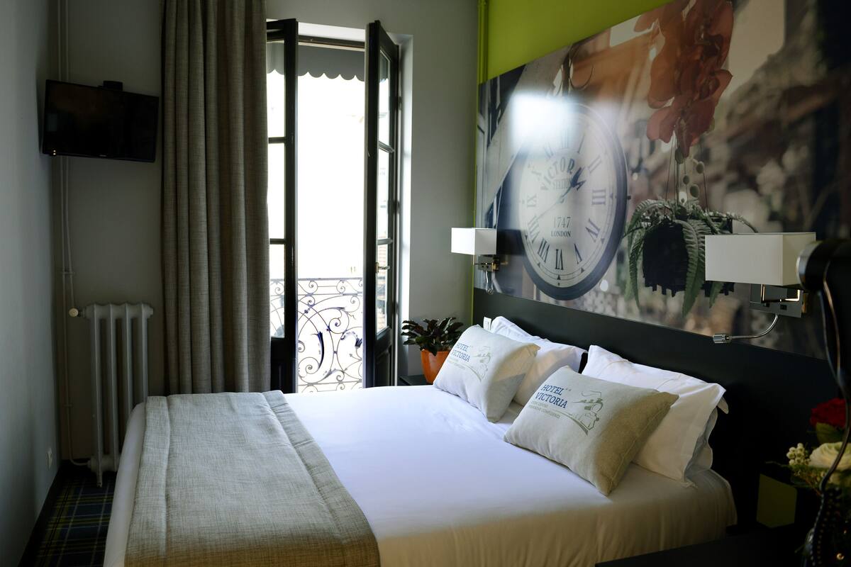 A hotel room with a bed, a television, a clock on the wall, and a window with curtains.