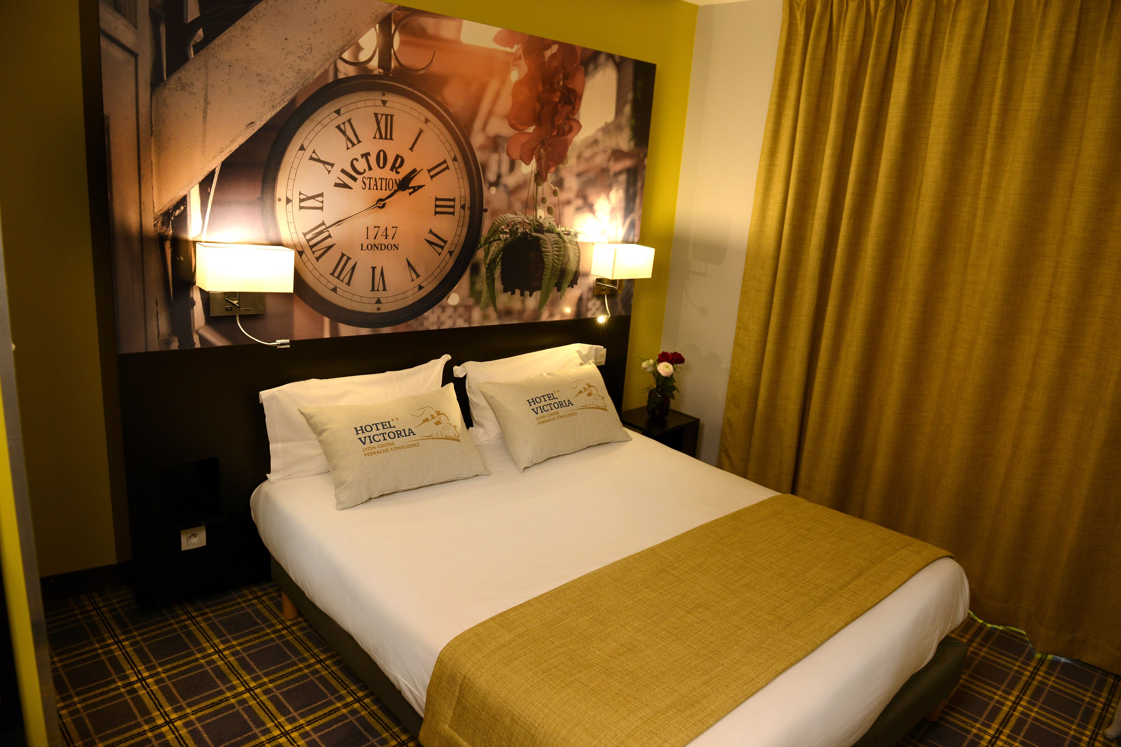 Une chambre d’hôtel avec une grande horloge murale, deux lampes de chevet et un lit recouvert d’un tapis à carreaux.