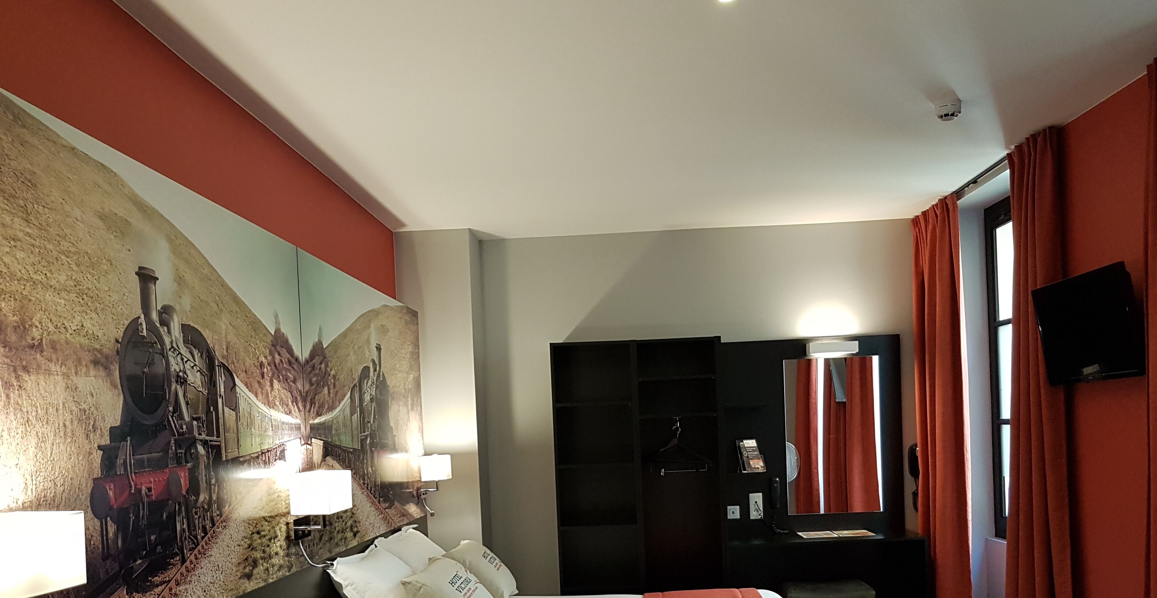 A hotel room with a bed, a large wall painting, a desk with a lamp, a mirror, and a TV.