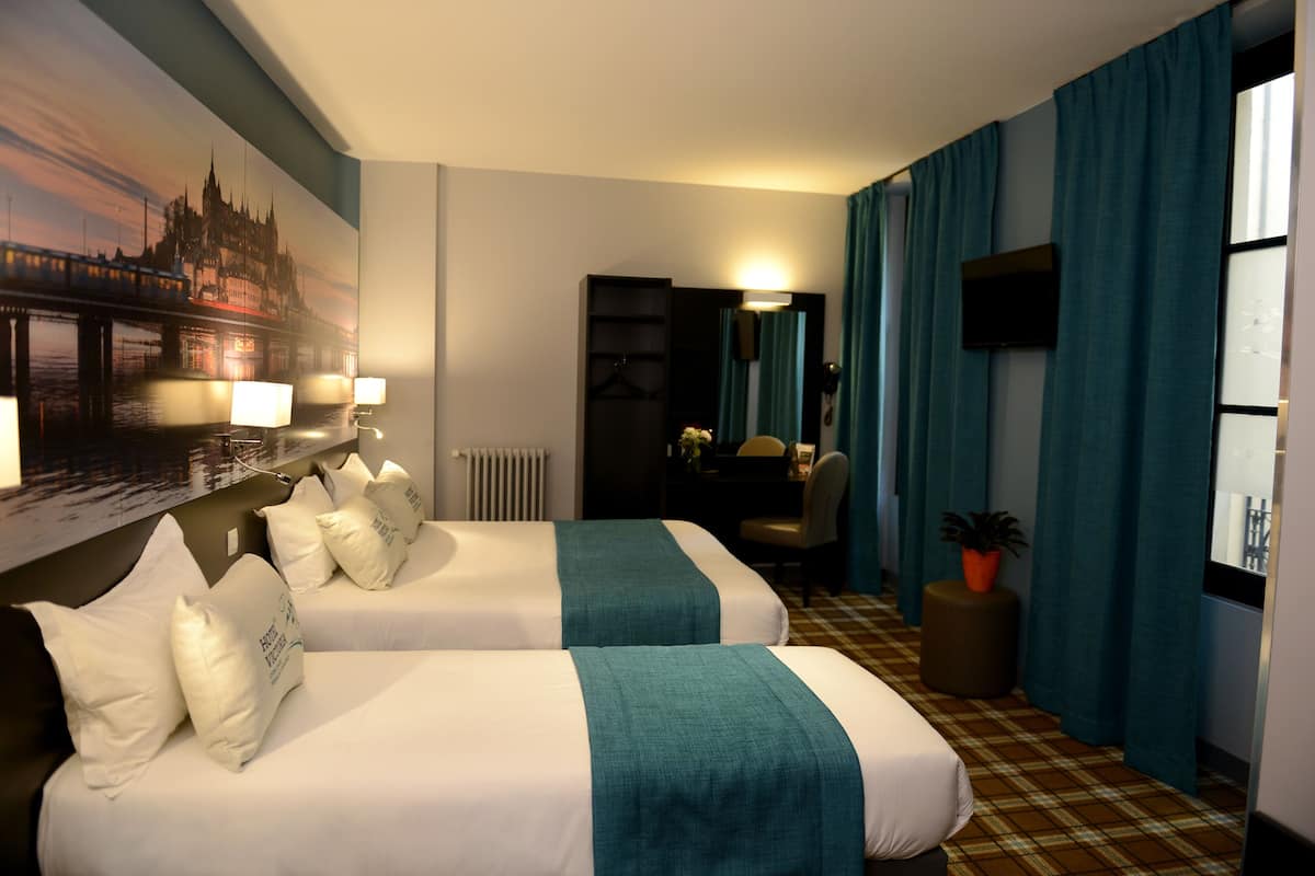 A hotel room with two beds, a large cityscape painting on the wall, a desk with a chair, a television, and a potted plant.