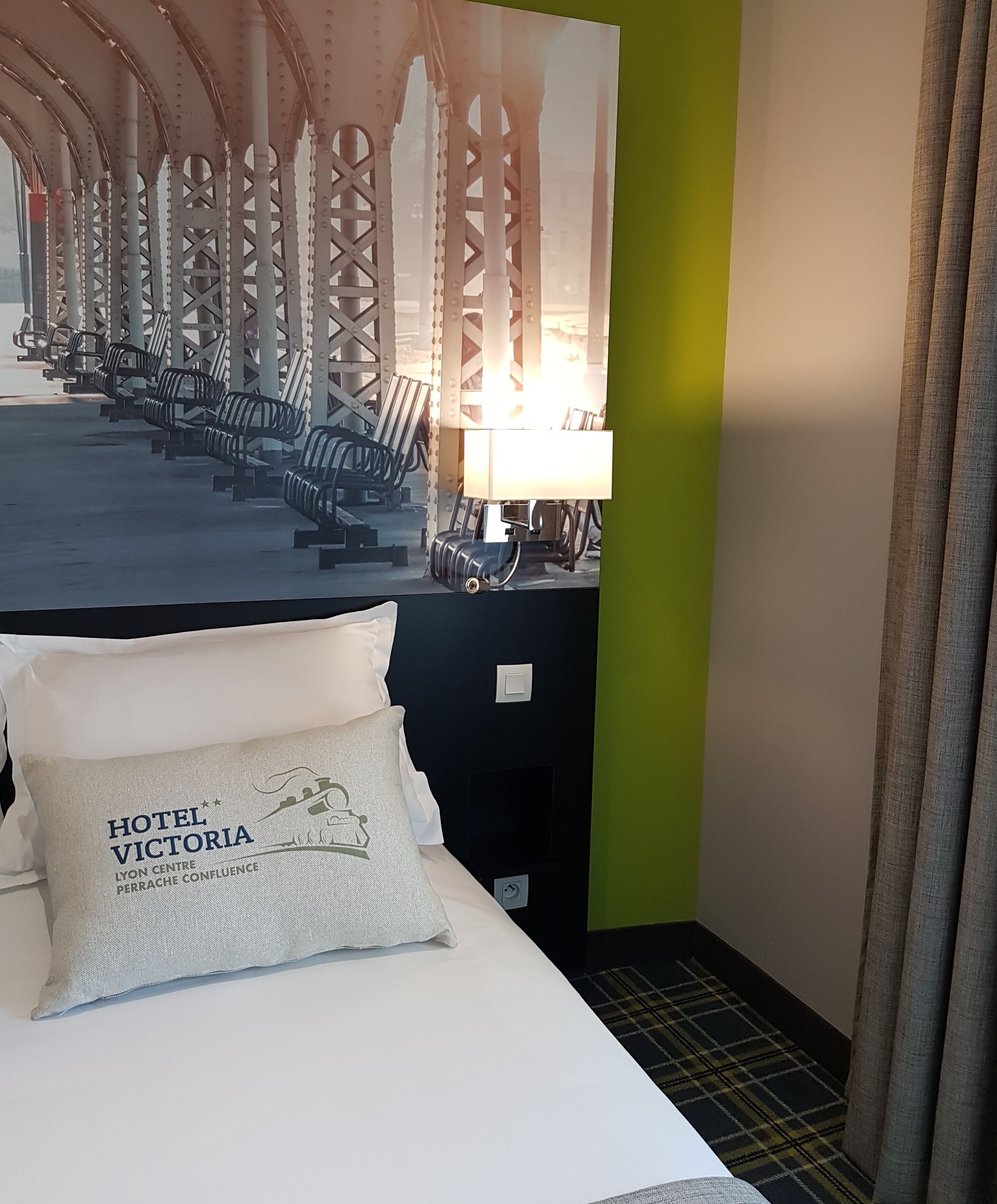 A hotel room with a bed, pillows, a nightstand with a lamp, and a wall with a bridge image.