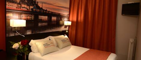 A hotel room with a bed, two pillows, a nightstand, a lamp, a vase with flowers, a painting of a cityscape, a television, and a window with curtains.