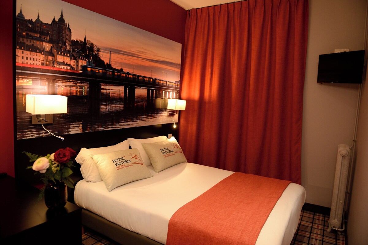 A hotel room with a bed, two pillows, a nightstand, a lamp, a vase with flowers, a painting of a cityscape, a television, and a window with curtains.