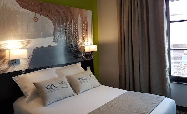 Habitación de hotel con cama, dos almohadas, mesita de noche, reloj de pared y ventana con cortinas.