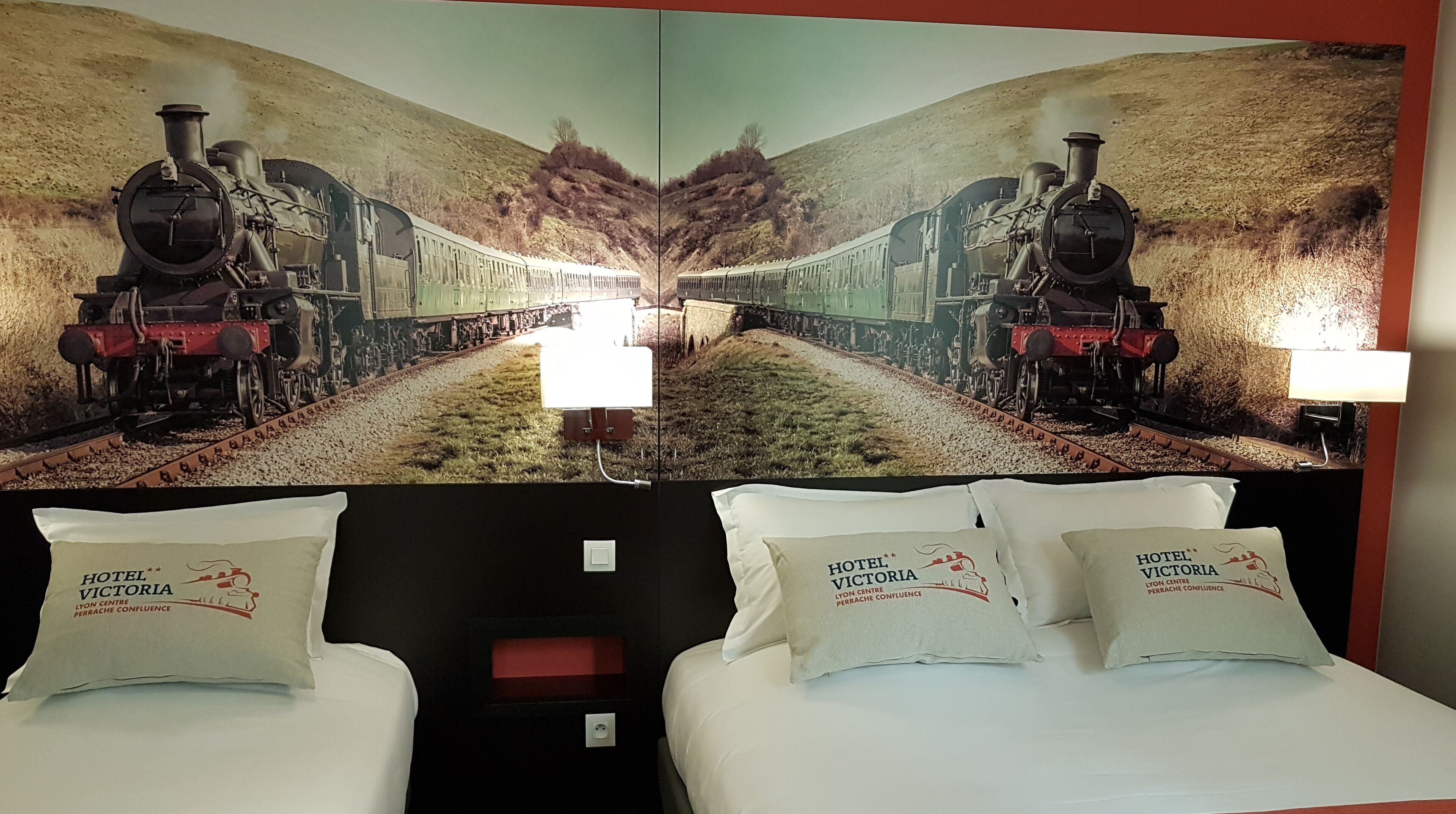 Habitación de hotel con pared espejada que muestra una imagen de tren, dos camas con almohadas y una pared de acento roja.