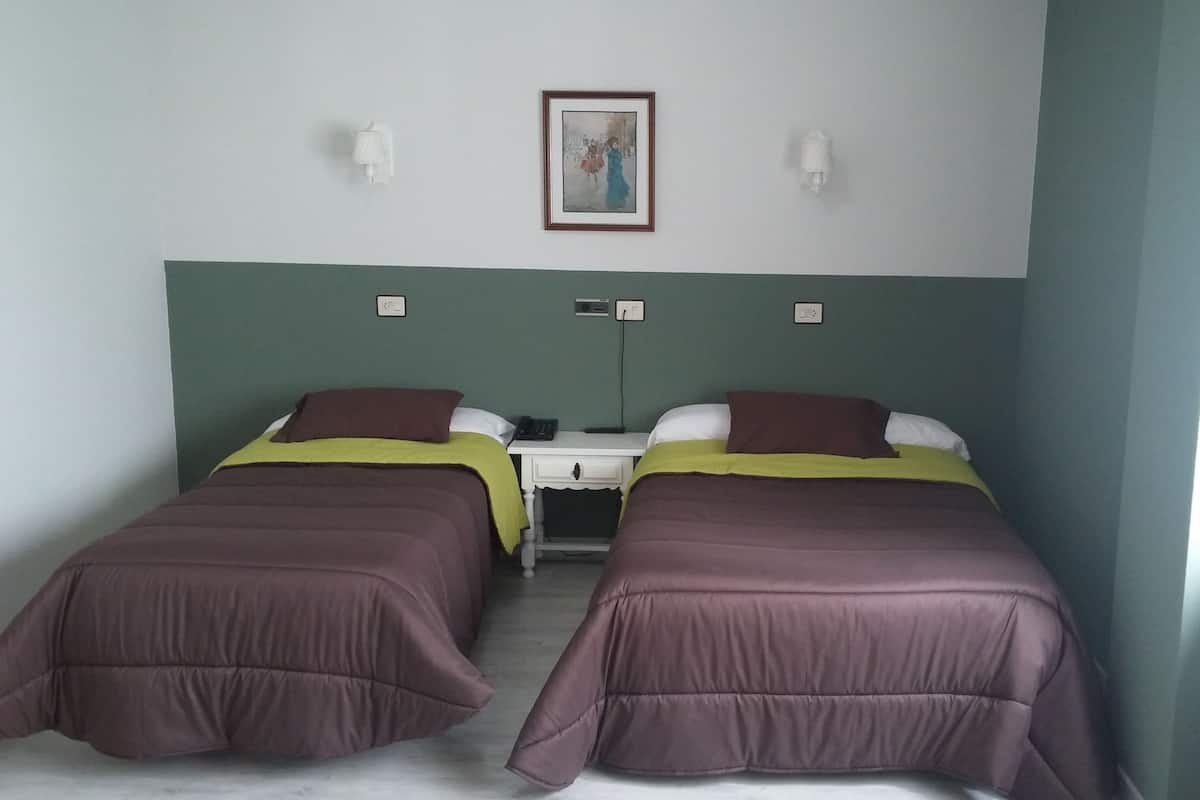 Une chambre double avec deux lits, chacun recouvert d’une couvre-lit violette et d’une couverture verte. Une petite table se trouve entre les lits, avec un téléphone et une prise électrique. Au-dessus de la table, un tableau encadré est accroché au mur.