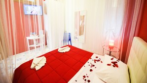 Habitación de hotel con una colcha roja, una mesita de noche blanca con una vela, y un televisor montado en la pared.