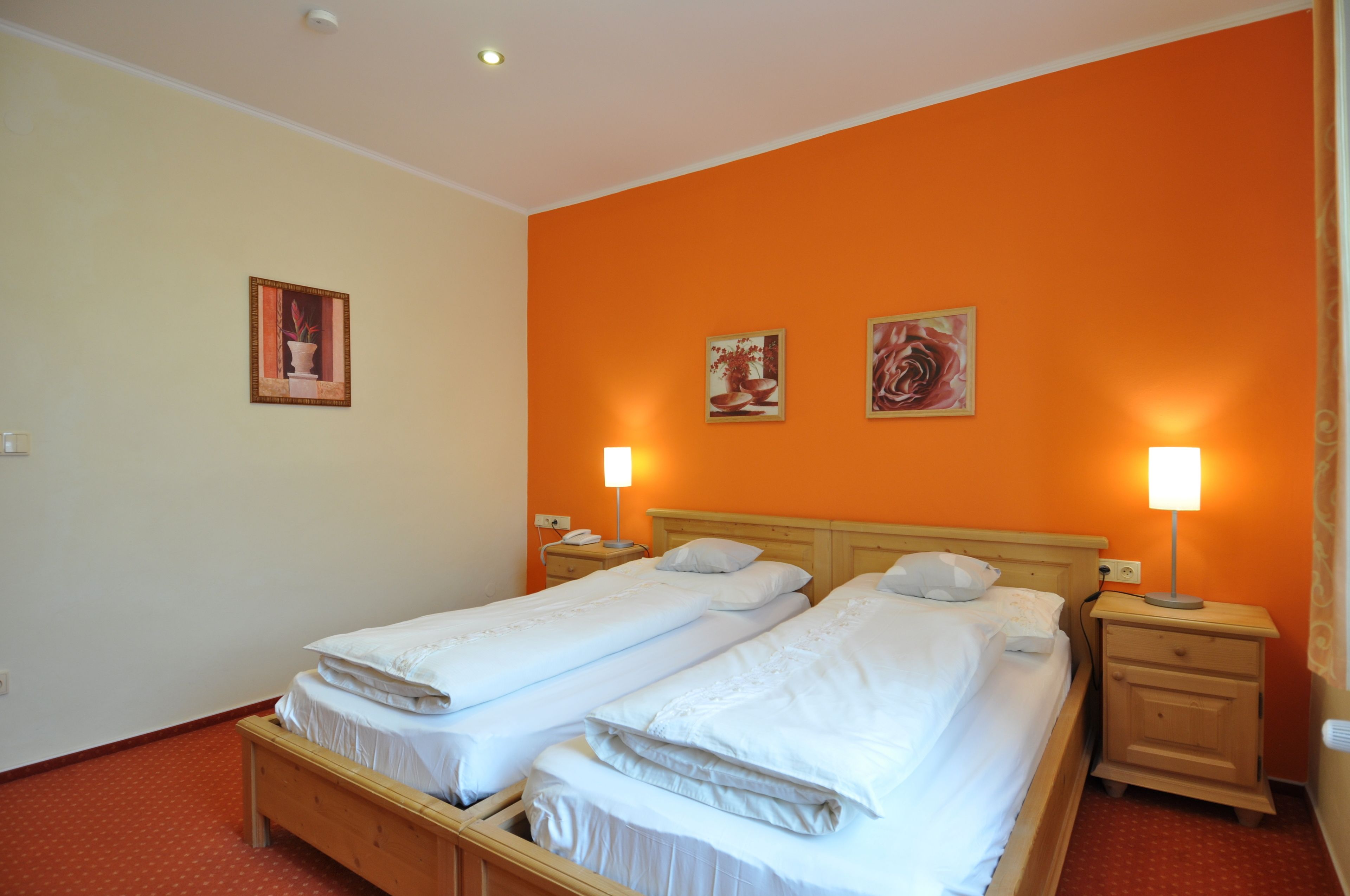 A hotel room with two single beds, a wooden headboard, bedside tables with lamps, and two framed pictures on the orange wall.