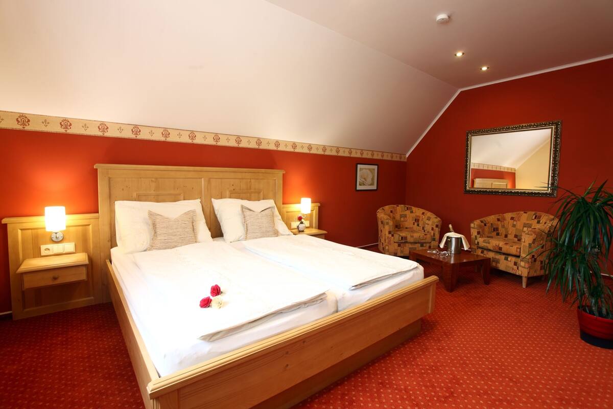A hotel room with a large bed, two bedside tables, a nightstand, a mirror, two armchairs, a potted plant, and a red carpeted floor.