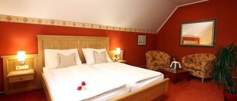 Habitación de hotel con una cama grande, dos mesitas de noche, un espejo, dos sillones, una planta en maceta y piso cubierto de alfombra roja.