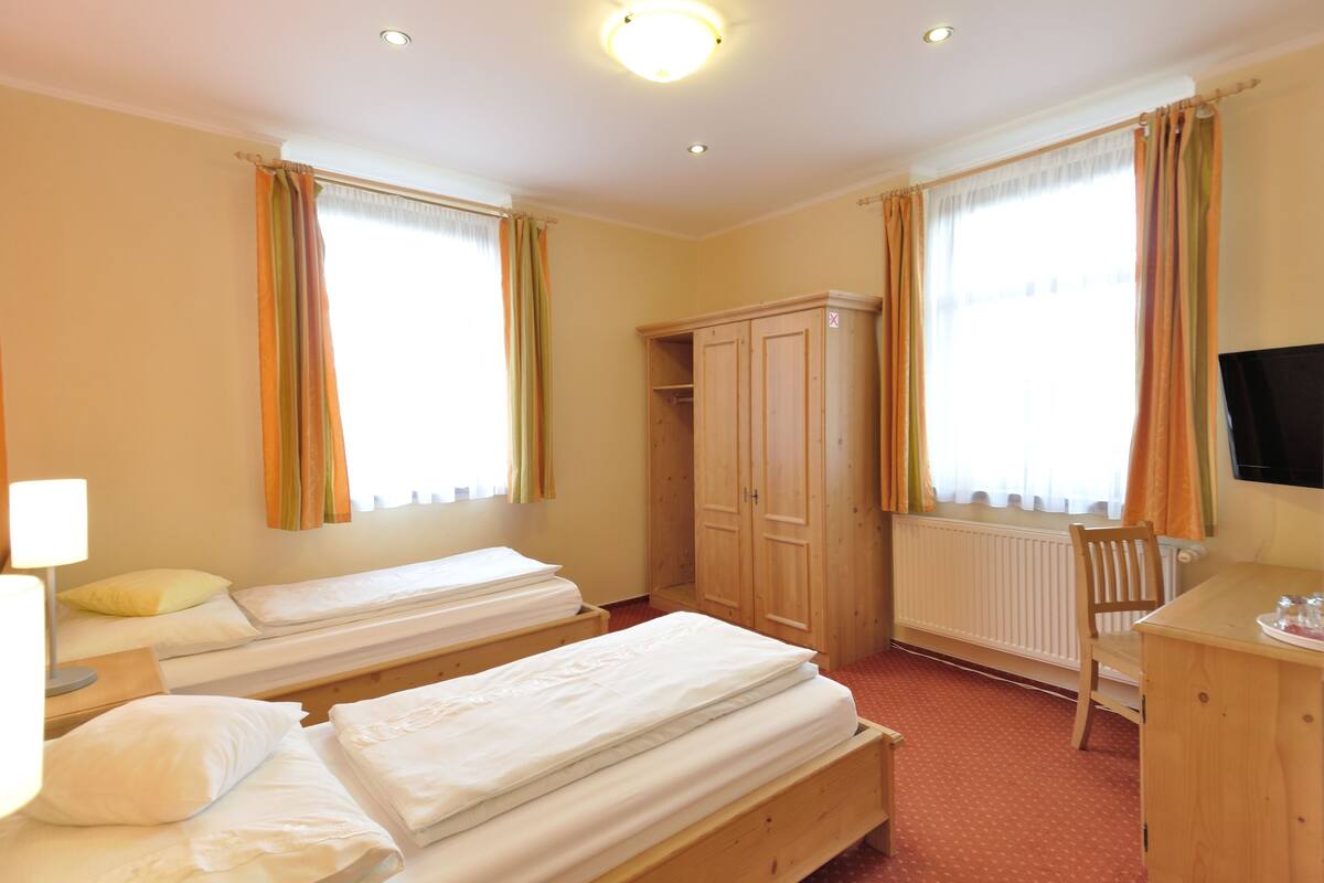 A hotel room with two single beds, a wooden wardrobe, a desk with a chair, and a television.