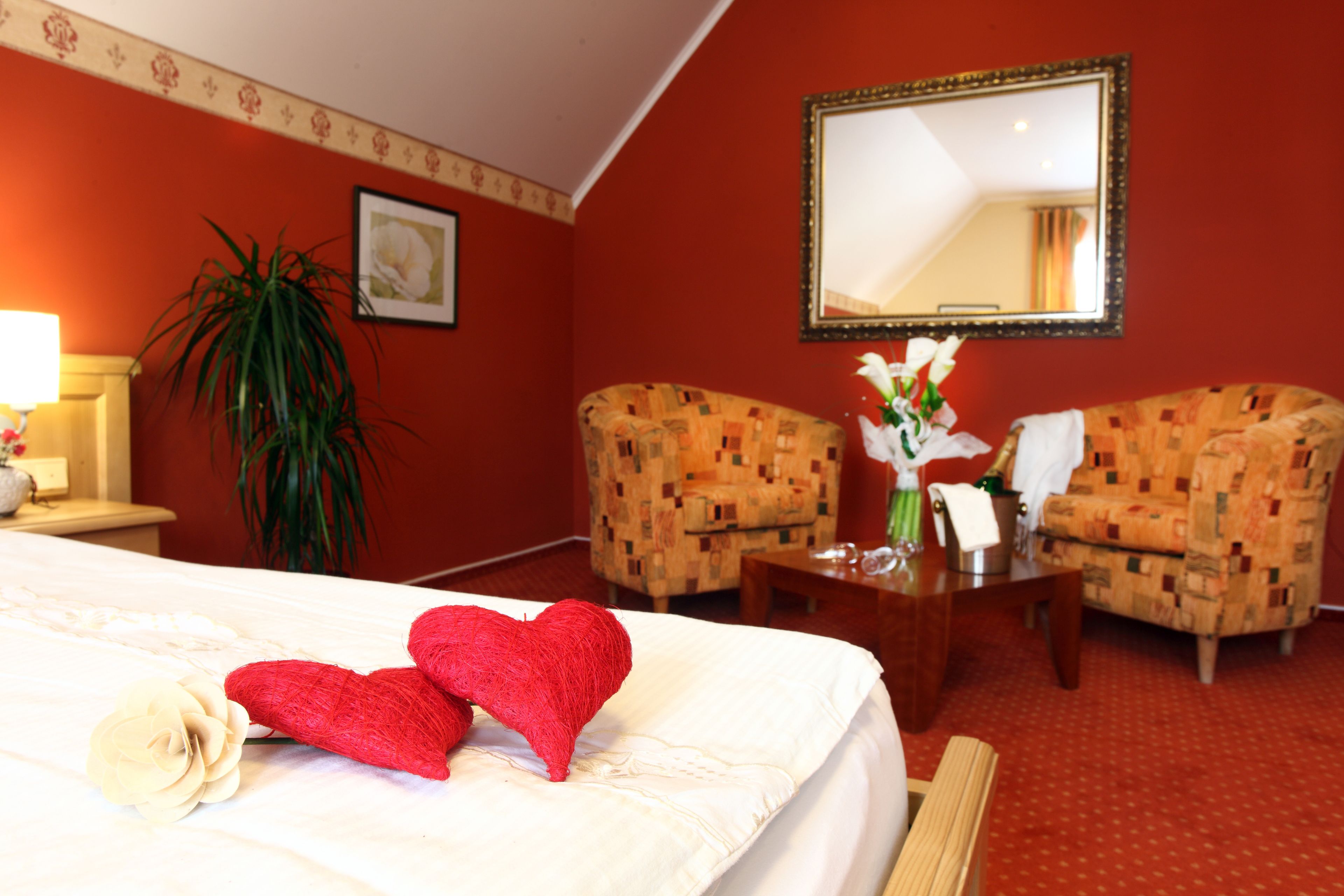 A hotel room with a bed, two armchairs, a small table with a vase of flowers, a mirror, and a potted plant.