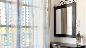 Una habitación con un escritorio de madera, un espejo, una silla y una ventana con cortinas translúcidas.