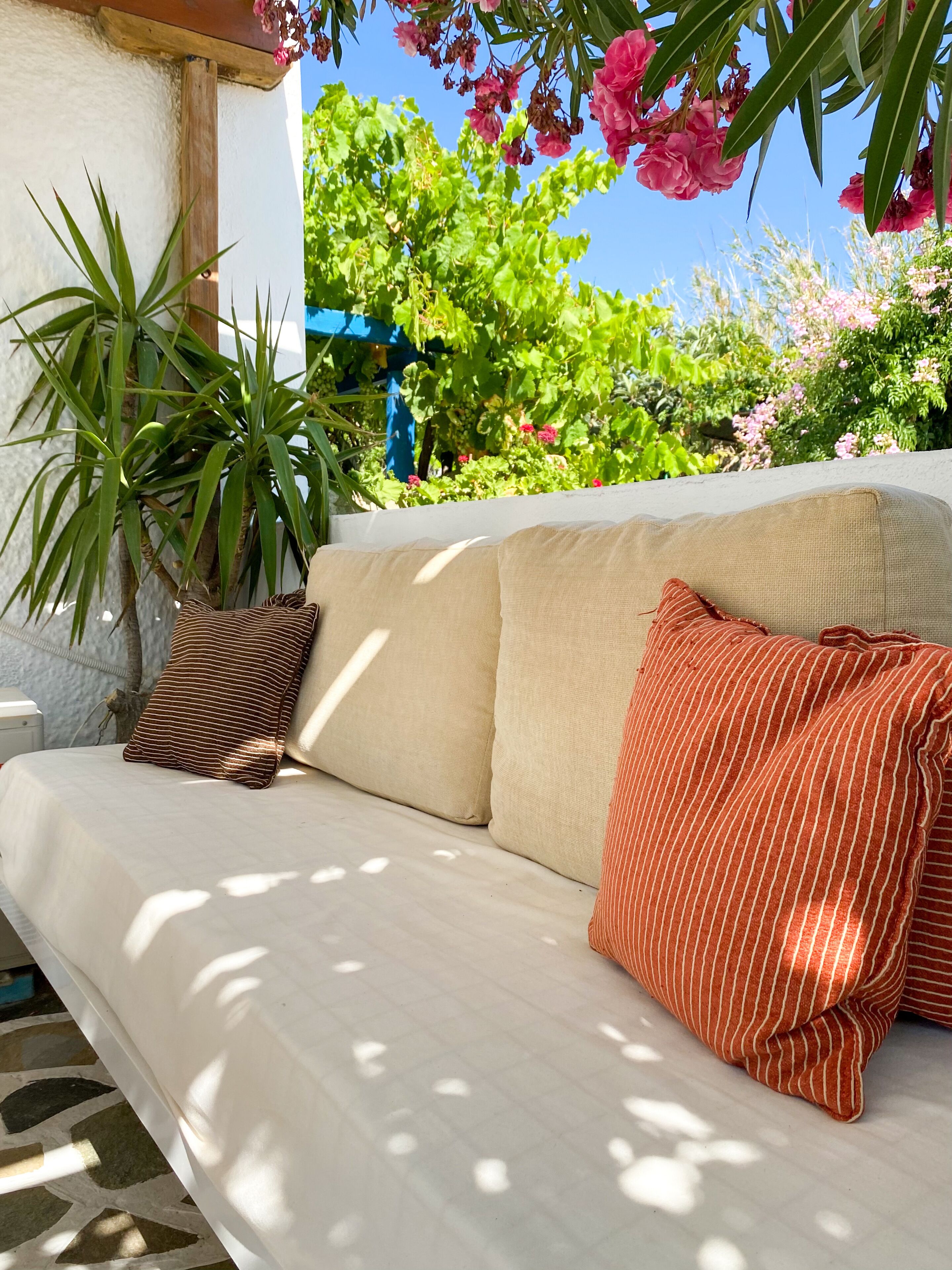Ein weißes Sofa mit gestreiften Kissen in einem sonnigen Außenbereich mit Pflanzen und Blumen.