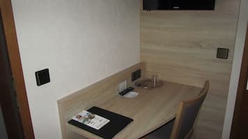 A small, modern hotel room with a wooden table, a chair, a TV mounted on the wall, and a small shelf with a glass and a remote control.