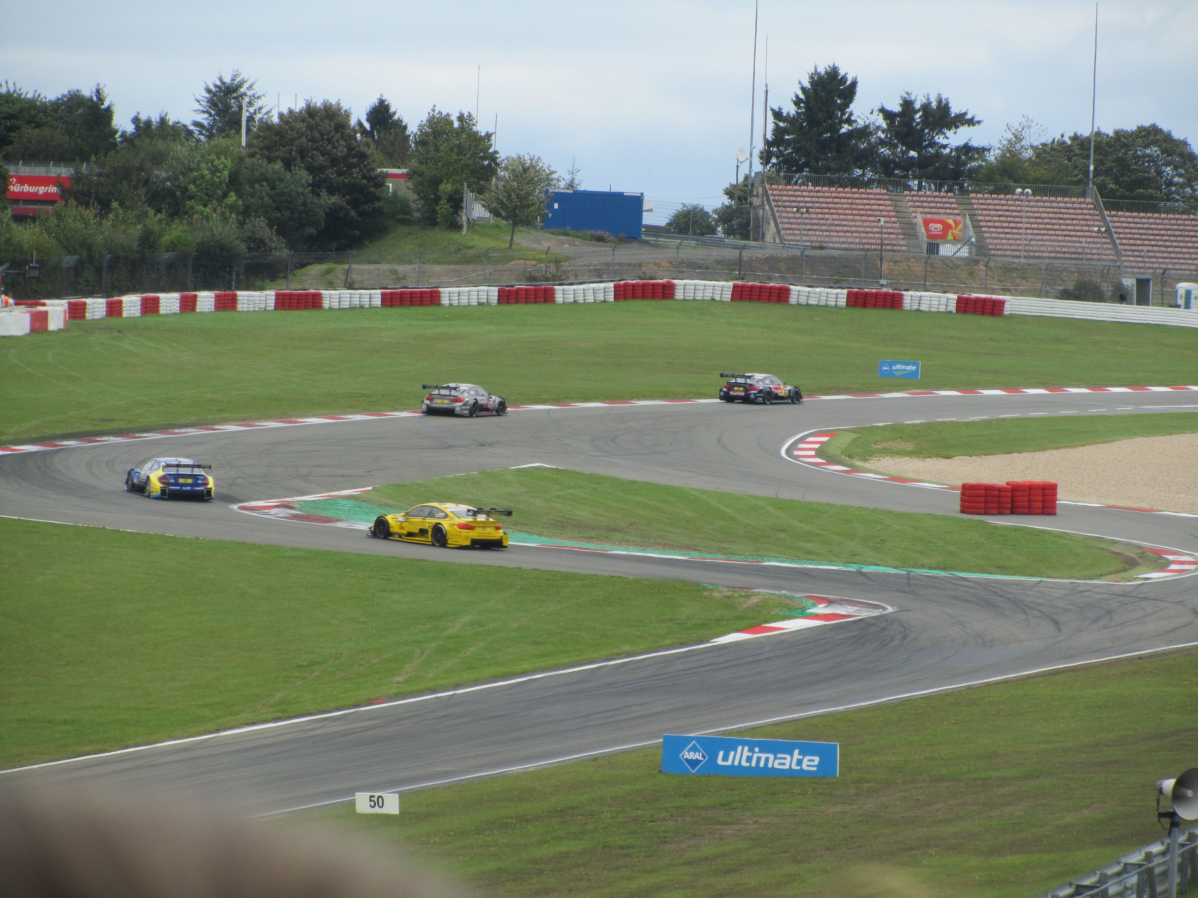 A race track with multiple cars in motion, surrounded by safety barriers and greenery.