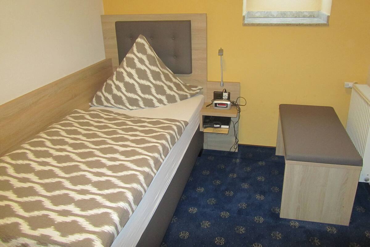 Ein kleines Zimmer mit einem Einzelbett, einer Bettdecke mit Muster, einem Nachttisch mit Lampe und einer Bank.