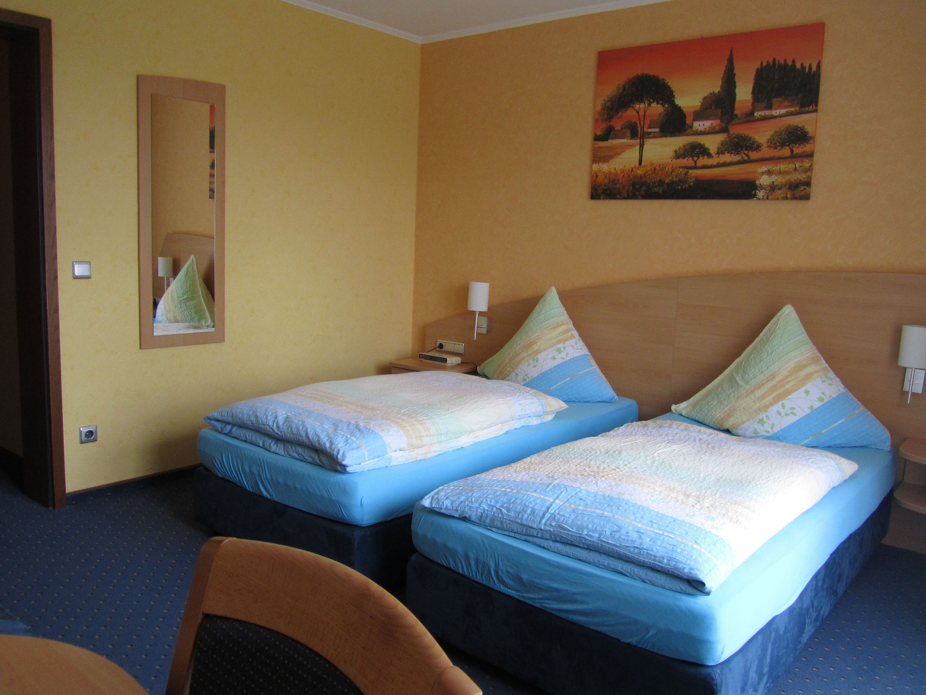 Una camera d'albergo con due letti, uno specchio, un quadro e un comodino con una lampada.