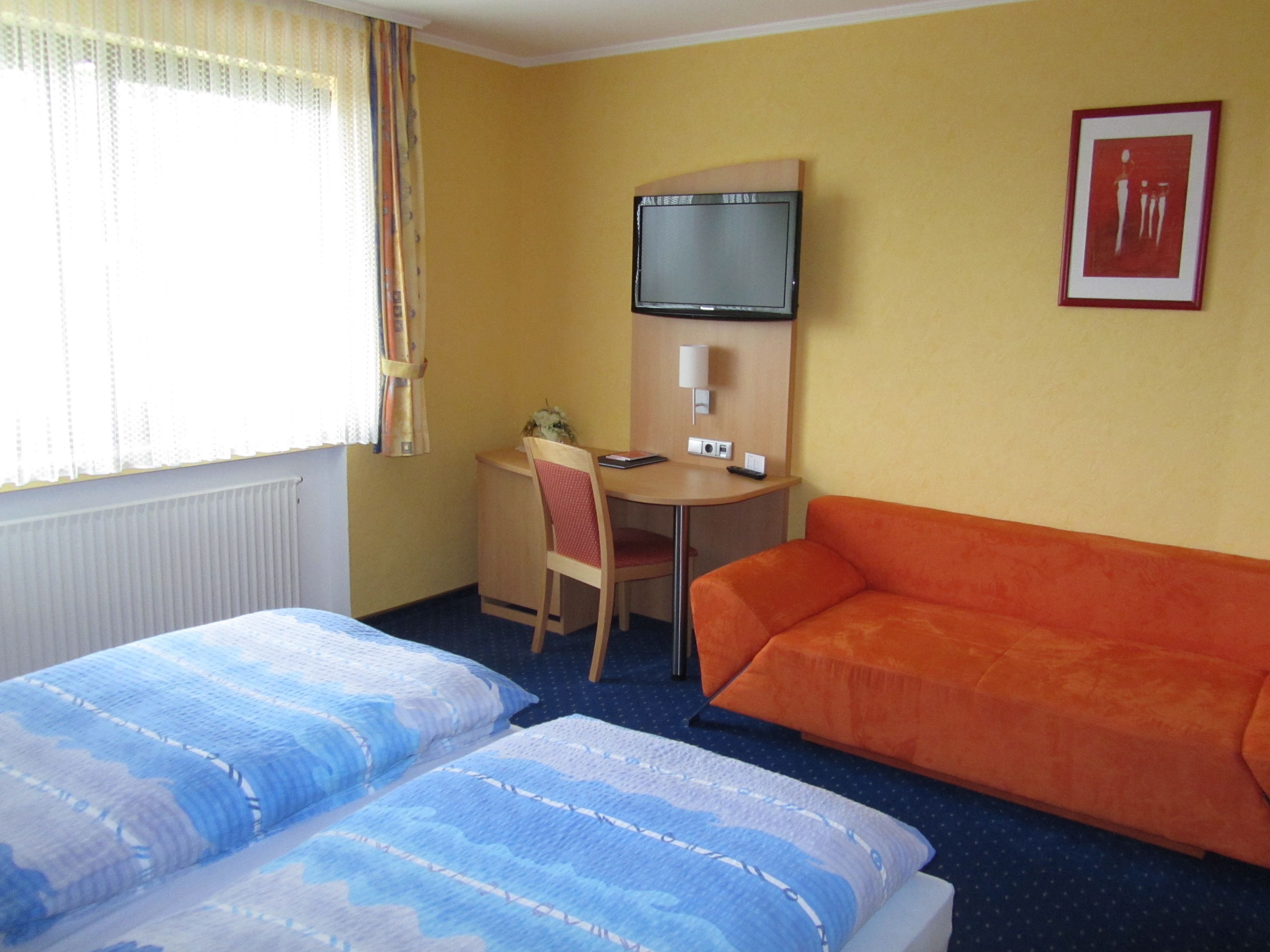 Een hotelkamer met twee bedden, een bureau, een stoel, een oranje bank, een televisie en een lijst aan de muur.