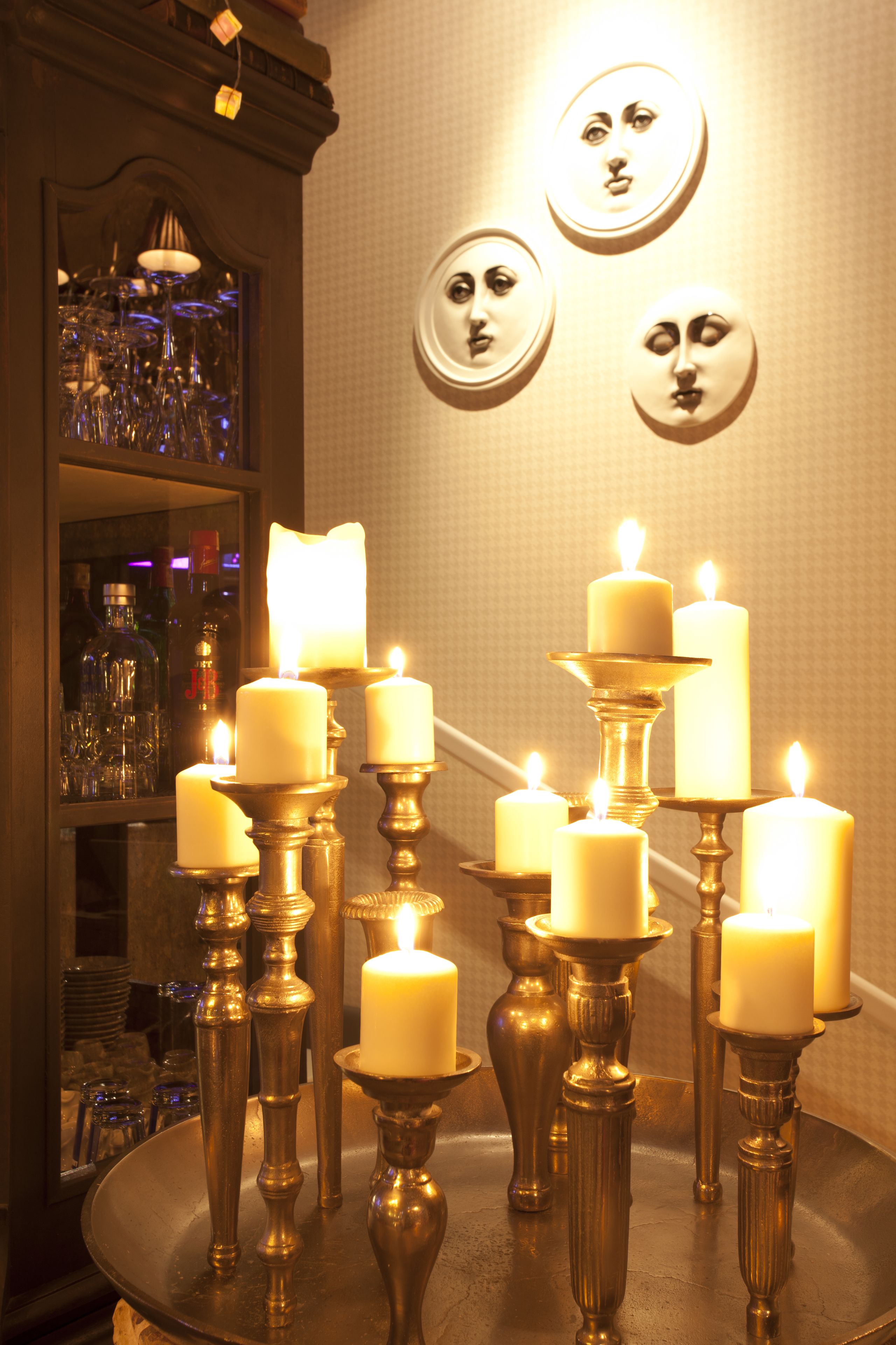 Um quarto com várias velas acesas em castiçais elaborados, um espelho refletindo um espaço de bar e pratos decorativos fixados na parede.