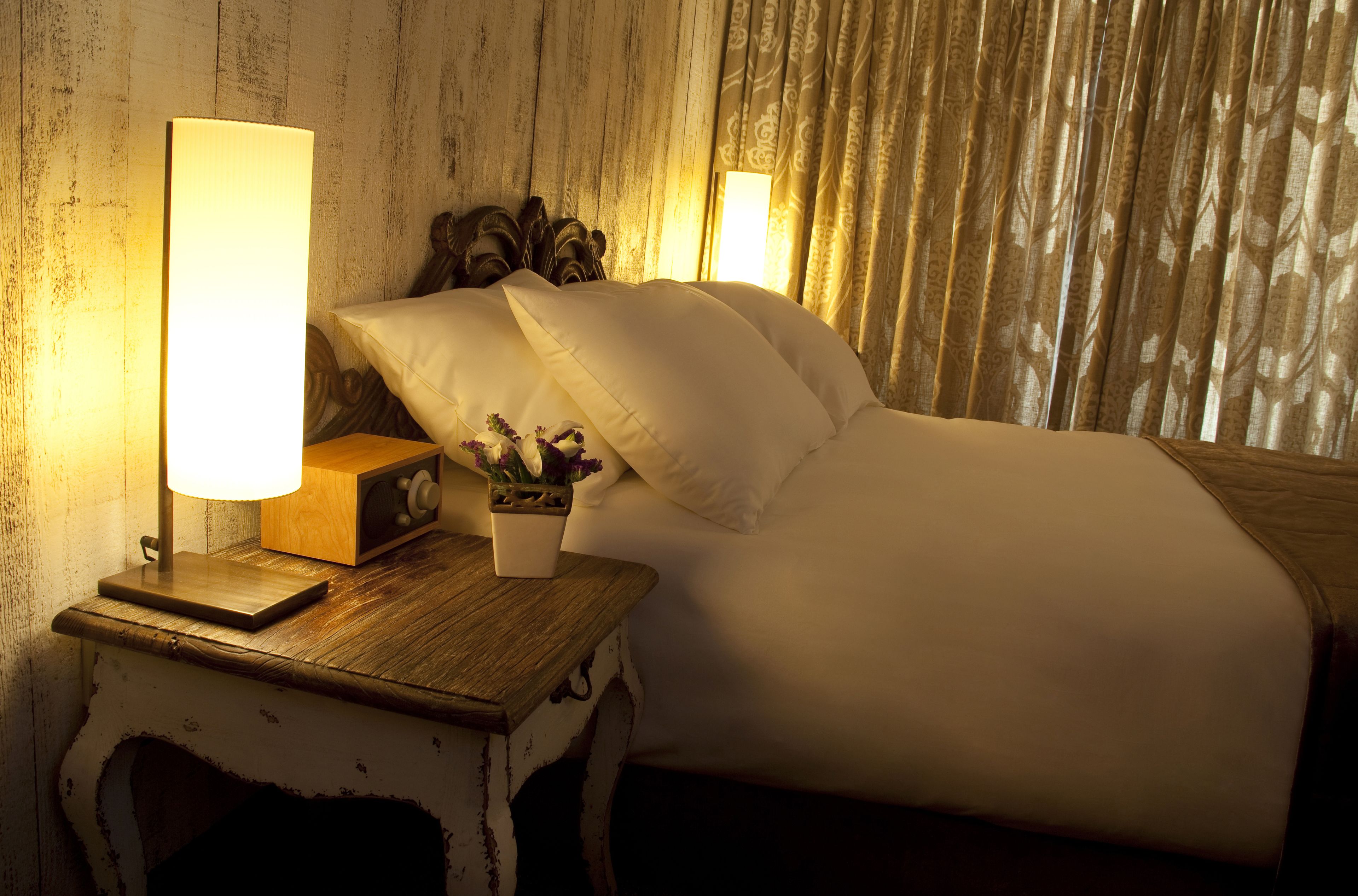 A neatly made bed with white linens, a bedside lamp, and a small wooden nightstand with a radio and a vase of flowers.