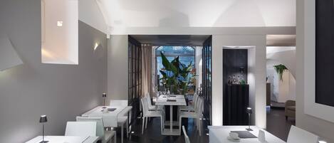 A modern dining area with white tables and chairs, a central plant, and a view through a glass door.