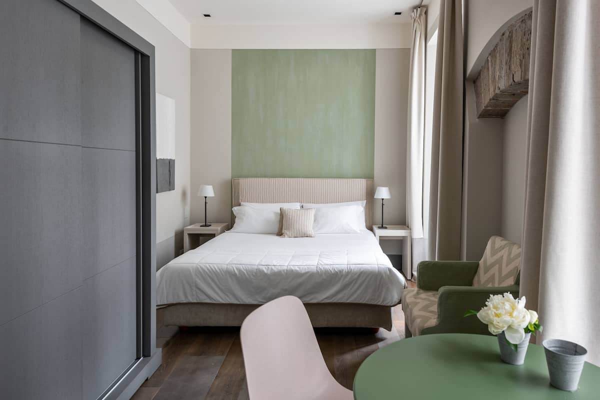 Una habitación de hotel moderna con una cama grande, dos mesitas de noche con lámparas, una silla verde y una pequeña mesa con un jarrón de flores.