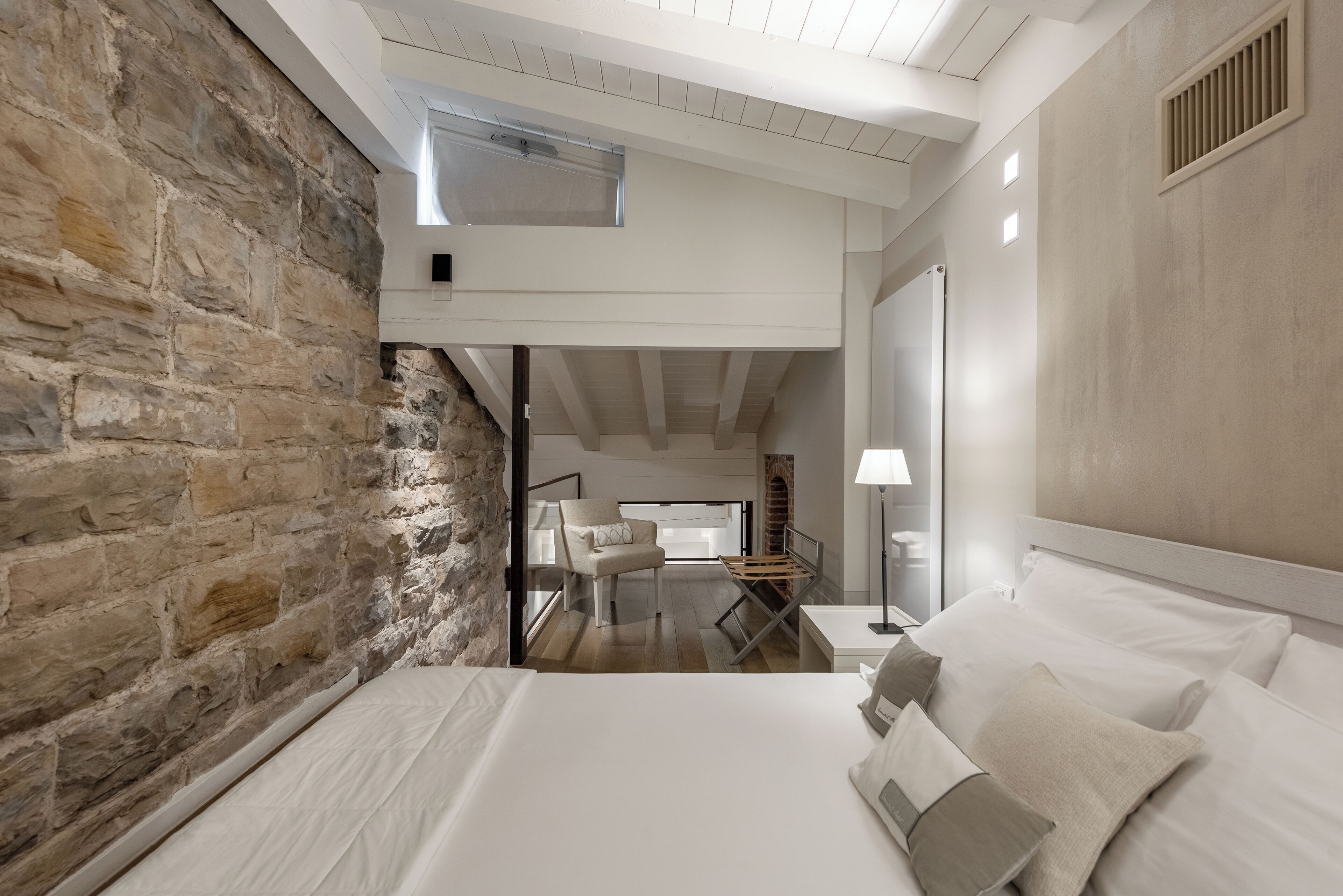 A bedroom with a stone wall, a bed with white linens, a chair, a small table, and a lamp.