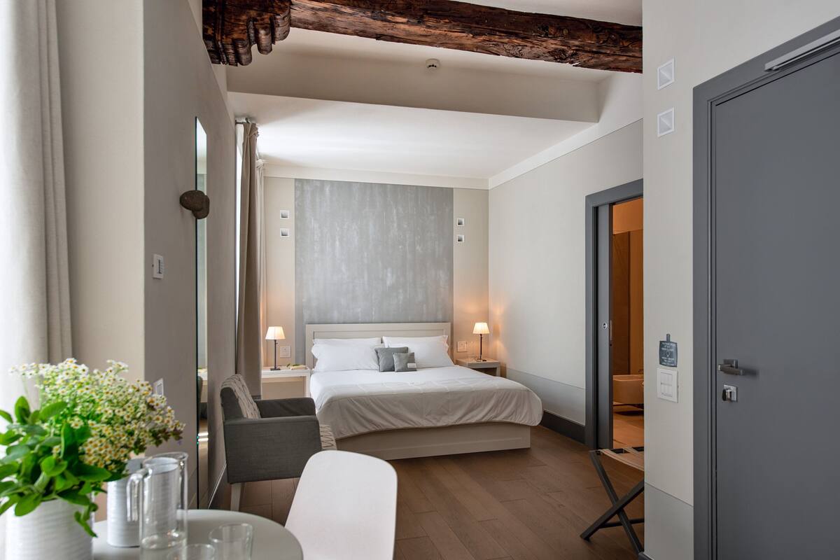 Una habitación de hotel moderna con una cama grande, mesitas de noche con lámparas, una pequeña mesa con un jarrón de flores y una puerta gris.