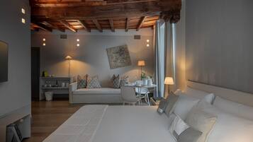A modern hotel room with a large bed, a sofa, a dining table, and a ceiling with exposed wooden beams.