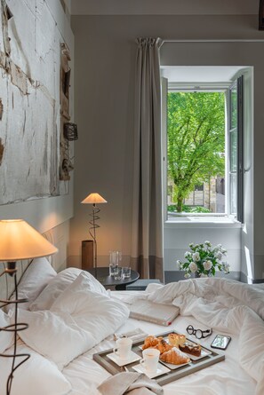 Hotelkamer met een bed, nachtkastje met lamp, dienblad met gebak en drankjes, een boek, en uitzicht op een boom door het raam.