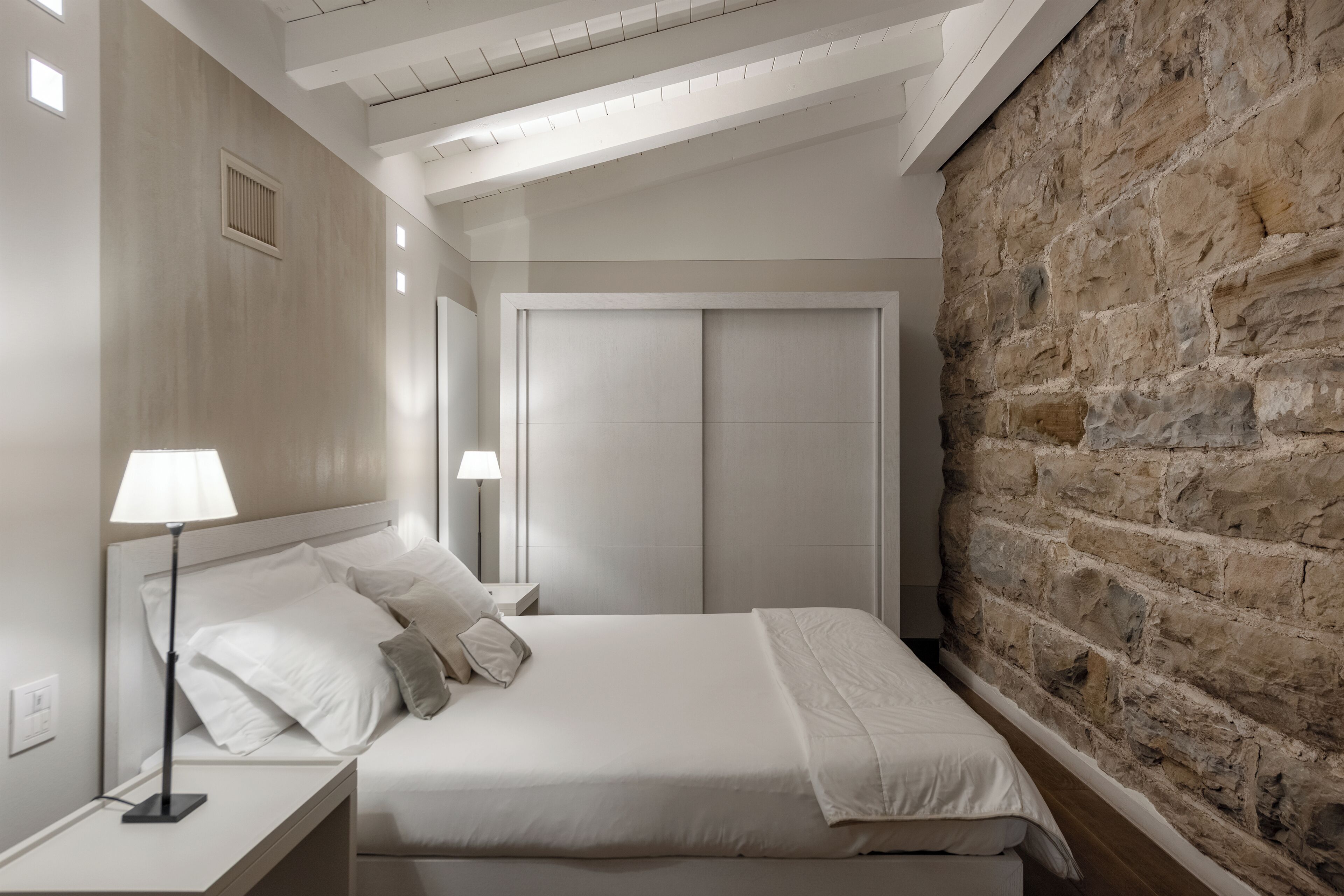 Un dormitorio con cama, lámparas de noche, armario y pared de piedra.