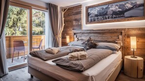 A bedroom with a large bed, a wooden headboard, a mounted picture of mountains, a small bedside table with a lamp, and a view of a balcony with chairs. - Sporthotel Romantic Plaza (Pinzolo)