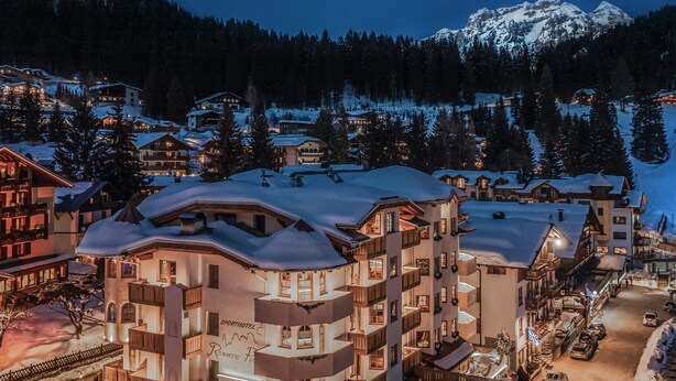A snow-covered mountain town at dusk with multi-story buildings, some with balconies and large windows, and a visible sign for a hotel named "Romantic Fl." - Sporthotel Romantic Plaza (Pinzolo)
