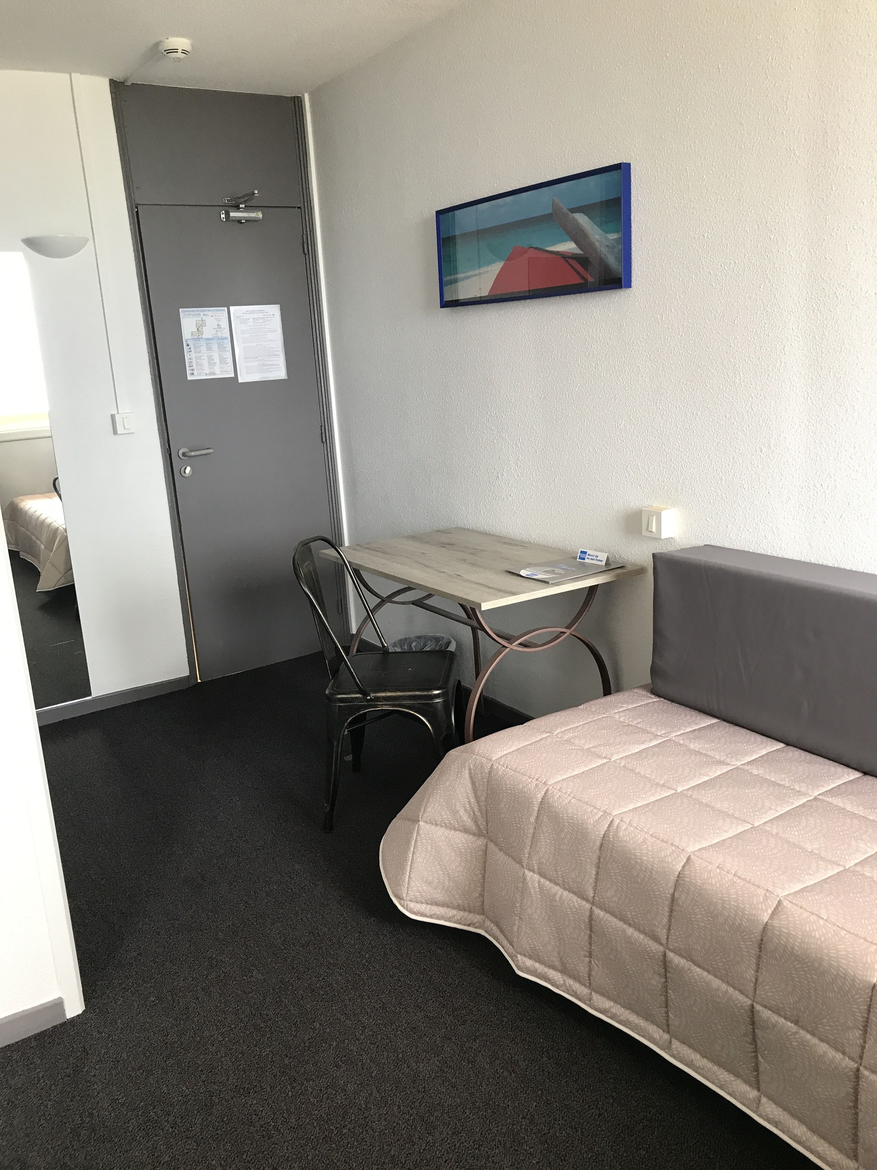 Hotelkamer met een bed, een bureau en een stoel. Er is een deur met een hendel en een lichtschakelaar. Een schilderij hangt aan de muur boven het bureau.