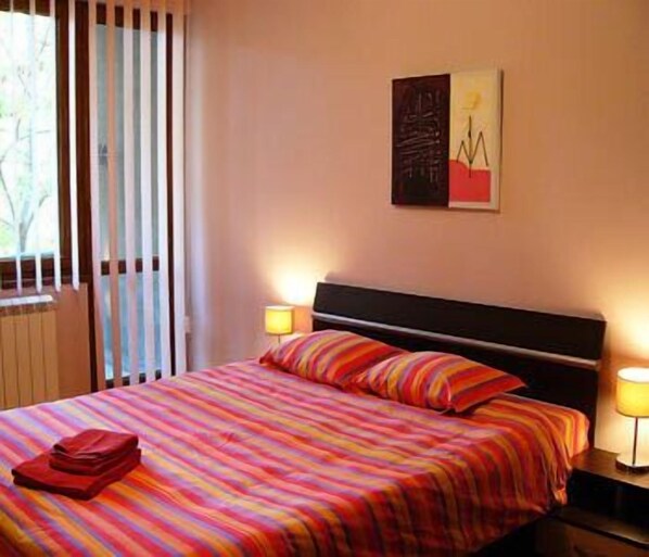 A hotel room with a bed, striped bedding, a nightstand with a lamp, and a window with blinds.