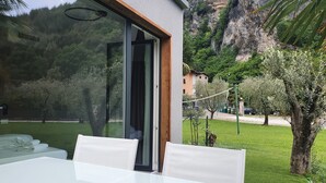 Un espacio moderno para comer al aire libre con un ventanal de vidrio que da a un jardín exuberante, rodeado de un paisaje montañoso.