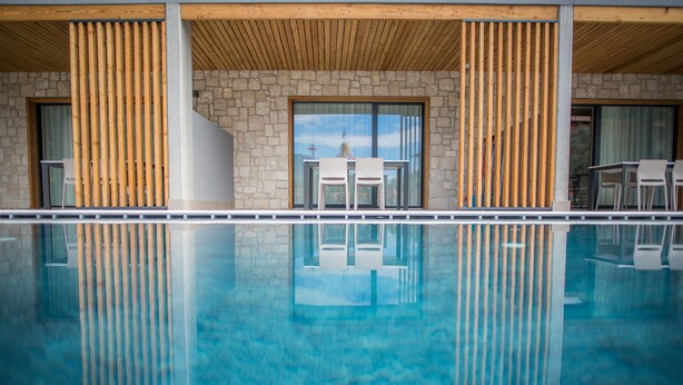 Zona de piscina moderna con pared de piedra y listones de madera, que refleja un cielo despejado y un edificio con puerta de cristal.
