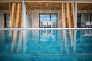 Un espace piscine moderne, agrémenté d’un mur en pierre et de lambris en bois, qui se reflète dans le ciel bleu et sur lequel se distingue un bâtiment doté d’une porte en verre.