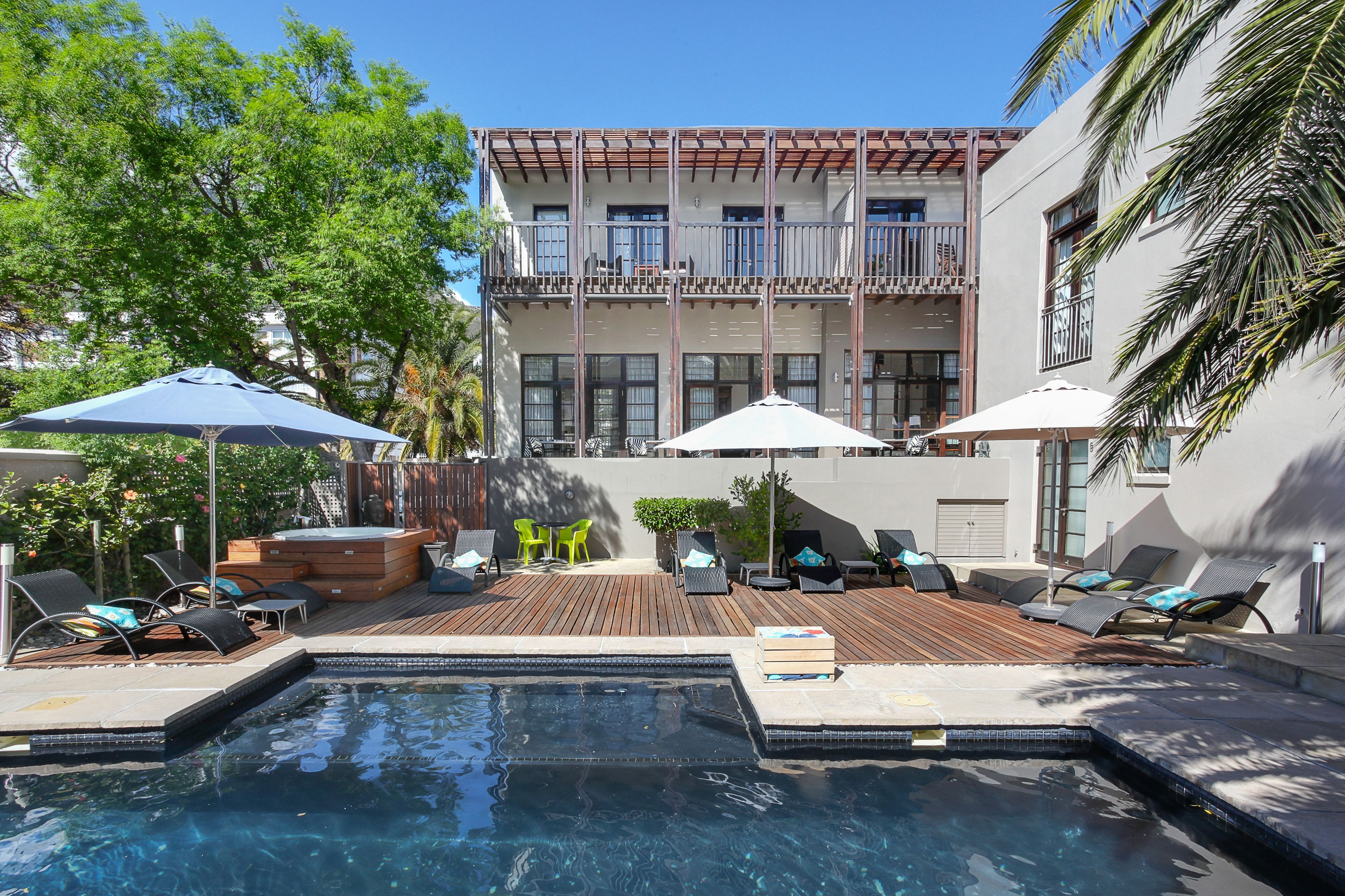 Un espace piscine avec des chaises longues et des parasols, une terrasse en bois et, au fond, un bâtiment doté de balcons.
