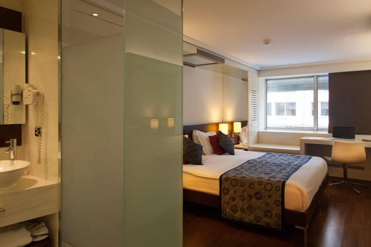 Ein modernes Hotelzimmer mit einem großen Bett, einem Nachttisch und einem Schreibtisch mit Stuhl. Es gibt ein Badezimmer mit Waschbecken und Spiegel.