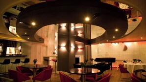 Zona pranzo moderna con tavoli rotondi e sedie rosse, un pianoforte e un bar sullo sfondo.