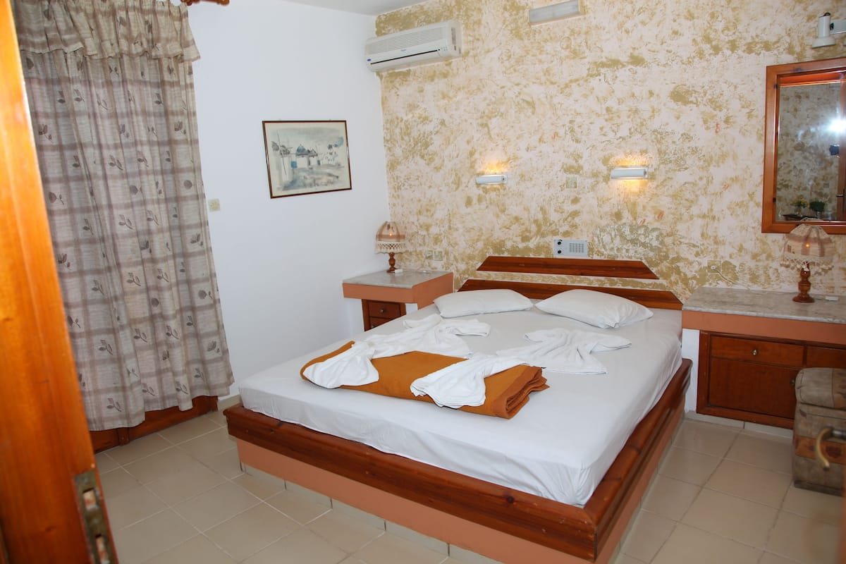 A bedroom with a wooden bed, a bedside table, a mirror, and a wall-mounted air conditioner.